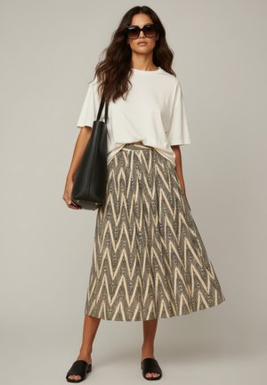 Vrouw draagt een oversized wit T-shirt, een midi rok met zigzag patroon, zwarte slidesandalen, een grote zwarte tote tas en vierkante zwarte zonnebril.