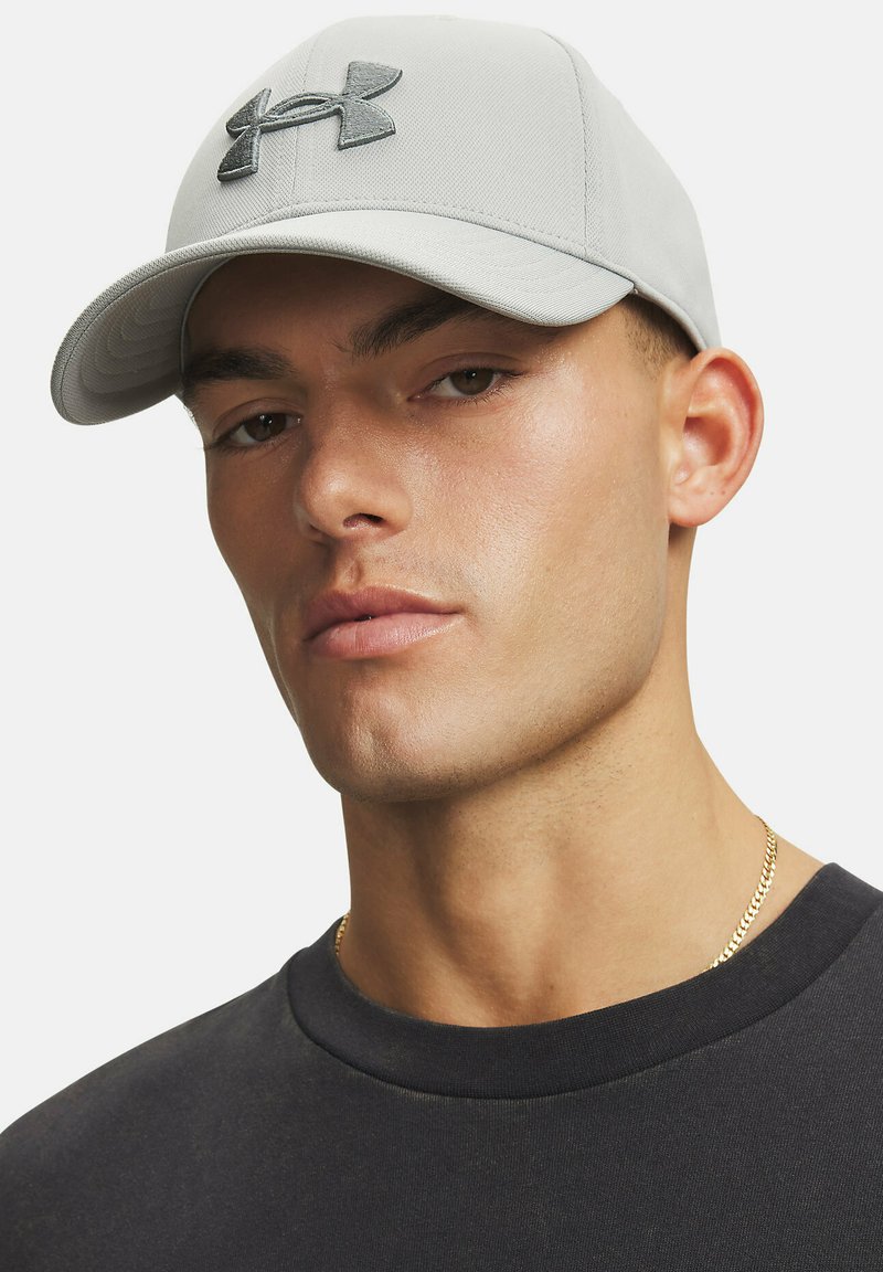 Cappellino grigio con una texture liscia e una visiera leggermente curva. Presenta un logo Under Armour grigio scuro ben visibile sulla parte anteriore.