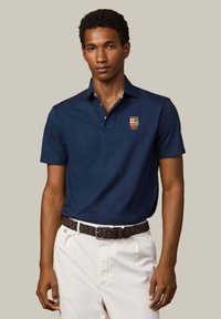 Hombre con una camisa tipo polo azul marino con un emblema de escudo y pantalones blancos, de pie contra un fondo liso beige.