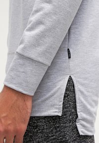 Graues Langarmshirt mit gerippten Manschetten, seitlichem Schlitz und einem Stoffetikett. Weicher Stoff, lässiges Design. Über dunkel gemusterten Hosen getragen.