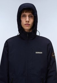 Giovane rivolto in avanti vestito con una giacca a cappuccio blu navy con il logo Napapijri Geographic sul petto e una toppa circolare sulla manica.
