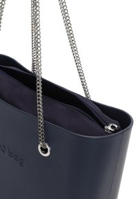 Tmavě modrá taška přes rameno s lesklým povrchem, stříbrnými řetízkovými popruhy, zipovým uzavíráním a vnitřní látkovou podšívkou. Na vnějším povrchu se nachází logo.