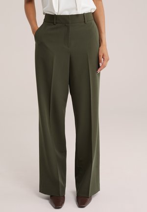 Femme portant un pantalon taille haute, large et vert olive avec poches latérales, et des chaussures marron pointues, associé à un chemisier blanc rentré.