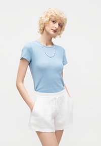 Νεαρή γυναίκα με κοντά σγουρά ξανθά μαλλιά που φοράει γαλάζιο κοντομάνικο τοπ, άσπρα σορτς με κορδόνι και κολιέ με χάντρες.