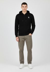 Felpa nera con zip, coulisse, tasche frontali e logo grafico bianco. Indossata con pantaloni grigi chiari e sneakers bianche, sfondo neutro.