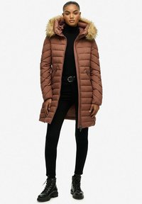 Brązowa kurtka puchowa z futrzanym kapturem, zapinana na zamek, o pikowanej strukturze. Nosi się ją na czarnym golfie i czarnych spodniach z butami na sznurowadła.