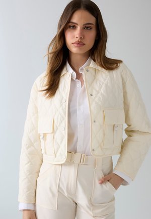 Młoda kobieta ubrana w kremową pikowaną kurtkę na białej koszuli zapinanej na guziki oraz kremowe spodnie z wysokim stanem i dopasowanym paskiem, ręka w kieszeni.