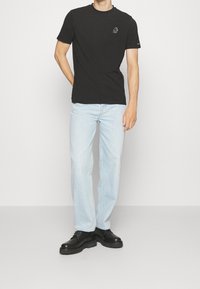 T-shirt nera in cotone con un piccolo logo sul petto, abbinata a jeans in denim blu chiaro e scarpe nere maestose. Outfit semplice e casual.