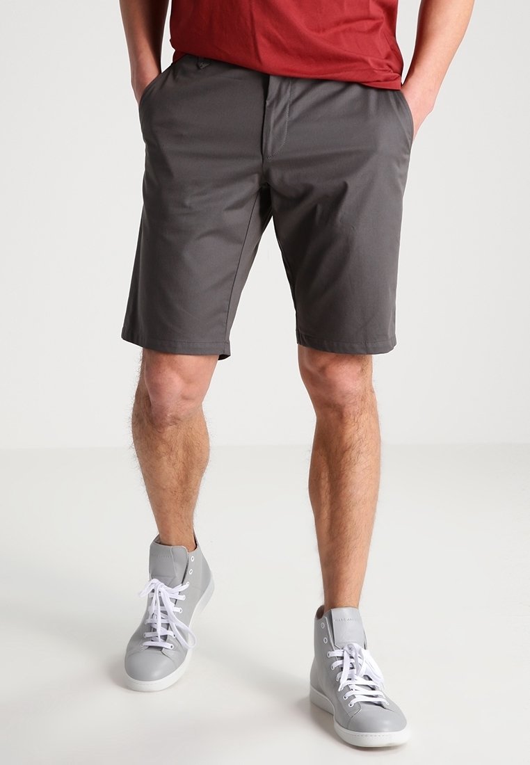 Homme portant un short gris foncé, une chemise rouge et des baskets montantes gris clair, debout les mains dans les poches.