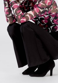 Top trasparente con motivi geometrici colorati in rosa, rosso e nero abbinato a pantaloni larghi neri e tacchi alti neri a punta.