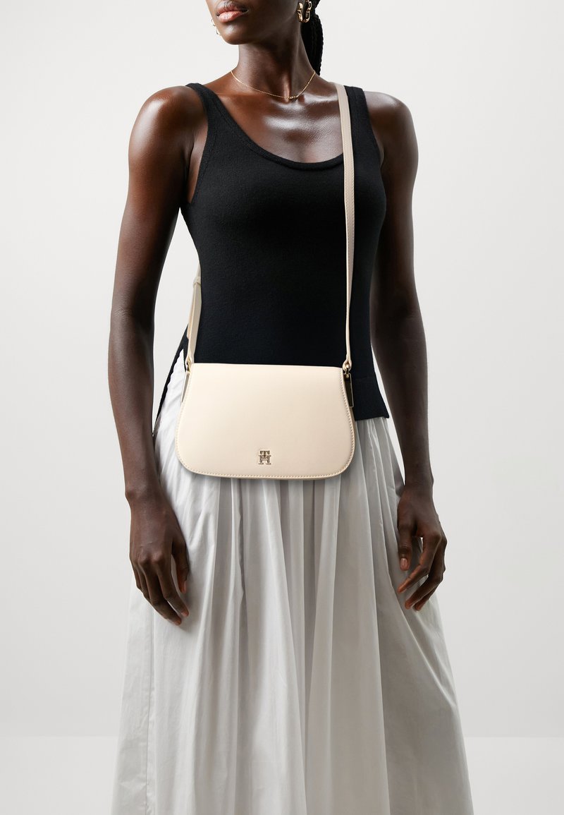 Cremfarbene Crossbody-Tasche mit geschwungener Form, goldfarbenen Beschlägen und geprägt Logo. Getragen über einem schwarzen Tanktop und gestuftem Rock.