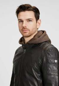 Chaqueta de cuero negro con un cuello alto, que presenta una capucha marrón debajo, detalles acolchados en los hombros y un parche con el logo en el lateral.