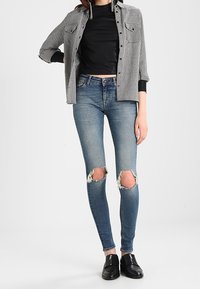 Kobieta w podartych niebieskich rurkach, czarnej bluzce, czarno-białej w kratkę kurtce i czarnych butach na zamek, stojąca na tle białym.