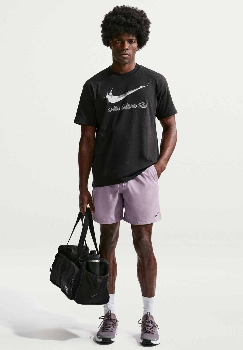 Homme avec coiffure afro portant un t-shirt noir Nike, un short violet clair, des chaussettes blanches, des baskets, tenant un sac de sport noir avec une bouteille d'eau.