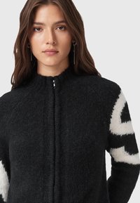 Maglione nero in lana con colletto alto, dotato di zip frontale e grandi motivi geometrici bianchi sulle maniche. Finitura morbida e testurizzata.