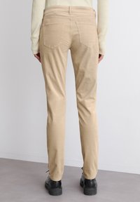 Beige puuvillapantsit, joissa on kapea malli ja takataskut, yhdistettynä mustiin kenkiin, nähtynä takaviistosta yksinkertaista taustaa vasten.