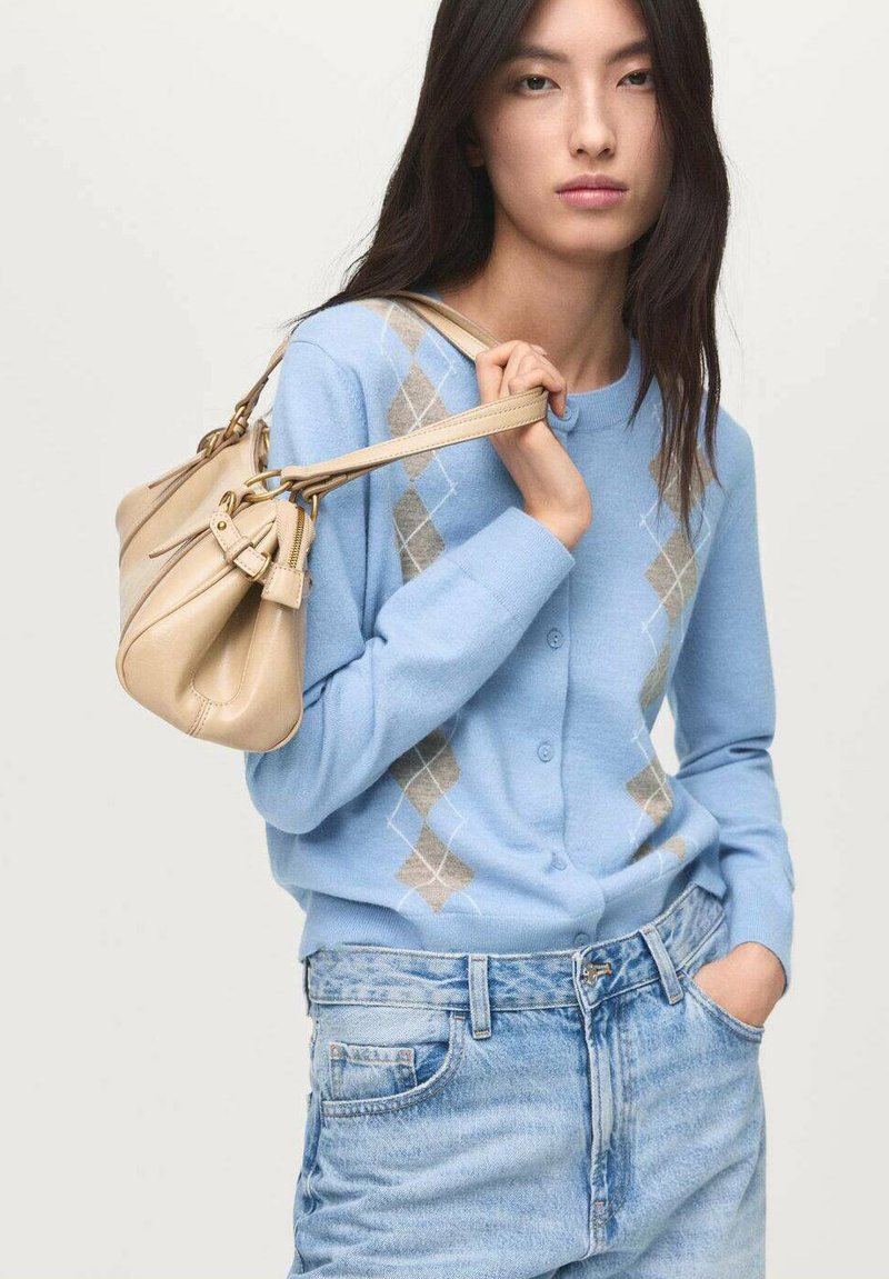Ljusblå cardigan med ett diamantmönster i beige och vitt, matchad med ljusa jeansshorts. Beigefärgad handväska med en strukturerad design.