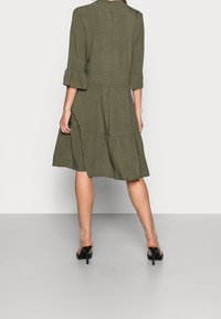 Žena má na sobě olivově zelené šaty ke kolenům s černými puntíky a volánkovými rukávy, doplněné černými lodičkami na vysokém podpatku.