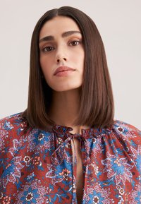 Blusa floreale rossa con motivi blu, rosa e bianchi. Presenta un collo arricciato e una chiusura a fiocco. Tessuto morbido e leggero.