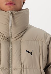 Giacca imbottita beige con colletto alto, texture a righe orizzontali e logo Puma nero sul petto. Visibile la cerniera frontale.