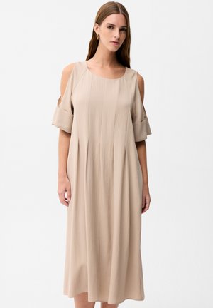 Kobieta ubrana w beżową sukienkę midi z odkrytymi ramionami, krótkimi rękawami i okrągłym dekoltem, stojąca na jednolitym białym tle.
