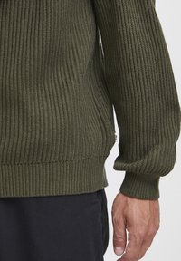Pulover din tricot ribbed de culoare verde măslin, cu un design texturat și ajustat, având mâneci cu manșetă și un mic etichetă pe cusătura laterală.
