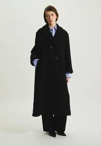Manteau noir croisé en tissu texturé, avec un grand col, des manches longues et des détails de boutons, s'étendant jusqu'au milieu du mollet.