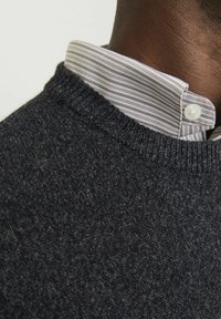 Suéter gris oscuro con un patrón de punto texturizado, combinado con una camisa de cuello blanca y azul claro a rayas visible en el escote.