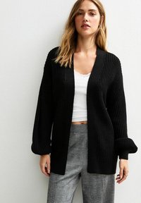 Cardigan nero lavorato a maglia con un motivo testurizzato, apertura frontale, maniche larghe e polsini a costine, abbinato a una maglietta bianca e pantaloni a quadri.