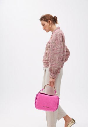 Une femme avec un chignon porte une veste rose texturée et un pantalon crème, tenant un sac à main rose vif avec une anse décorative.
