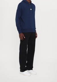 Marineblauer Baumwoll-Hoodie mit einer Fronttasche, verstellbarem Kordelzug und Logo-Patch. Kombiniert mit schwarzen Hosen und weißen Turnschuhen.