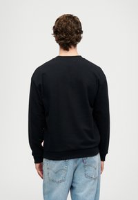 Felpa nera a collo rotondo con una texture morbida, caratterizzata da polsini e orlo a costine, abbinata a jeans in denim azzurro chiaro. Dettaglio della tag rossa sul lato.