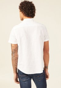 Camicia di lino bianca a maniche corte con colletto button-down, orlo curvato e maniche arrotolate, abbinata a jeans in denim scuro. Superficie testurizzata visibile.