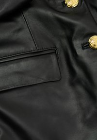 Czarna skórzana kieszeń kurtki z dwoma ozdobnymi złotymi guzikami na teksturowanym materiale z widocznym przeszyciem i detalami szwów.