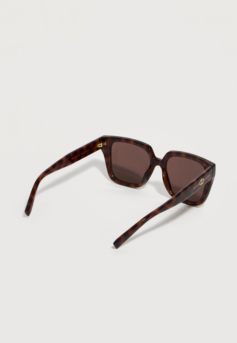 Tortoiseshell-patterned square sunglasses with dark lenses and gold logo on temple arms on a light background.