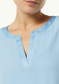 Blusa blu chiaro in tessuto morbido e liscio, con scollatura a V e dettagli sottili al colletto. Nessun motivo o accessorio visibile.