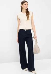 Donna che indossa un gilet crema senza maniche con bottoni, jeans ampi e scuri, scarpe crema, tiene una borsa beige, in piedi davanti a uno sfondo bianco.