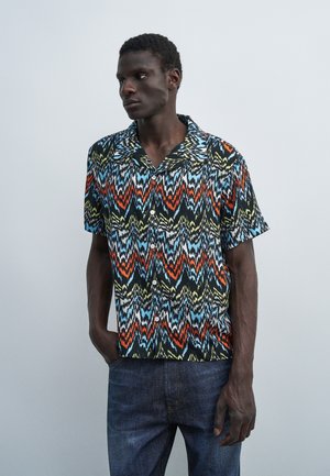 Homme portant une chemise à manches courtes à motifs zigzag multicolores et un jean bleu foncé, debout devant un fond gris clair uni.