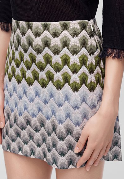 Gros plan d'une femme portant une jupe à motif zigzag multicolore avec des tons verts, bleus et beiges, associée à un haut noir texturé.
