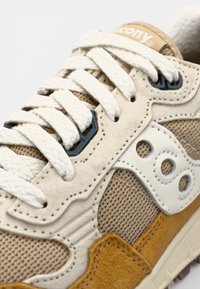 Beige sneaker with mesh and suede materials. Features a cream midsole, white leather accents, and thick white laces. Textured design details.