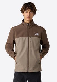 Ruskea ja beige pehmeäkuorinen takki korkealla kauluksella, vetoketjulla edessä ja sivutaskuilla. Ominaisuuksiin kuuluu The North Face -logo vasemmassa rinnassa.