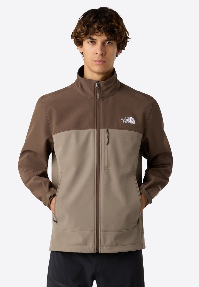 Ruskea ja beige pehmeäkuorinen takki korkealla kauluksella, vetoketjulla edessä ja sivutaskuilla. Ominaisuuksiin kuuluu The North Face -logo vasemmassa rinnassa.
