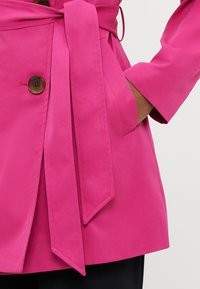 Ροζ παλτό με ζώνη, με πλαϊνή τσέπη, ένα μεγάλο καφέ κουμπί και λεία υφή με μακριά μανίκια, που αναδεικνύει μια ραμμένη σιλουέτα.