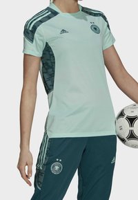 Osoba v jasně zeleném a tmavě modrém fotbalovém dresu Adidas drží bílou a černou fotbalovou kouli Adidas.