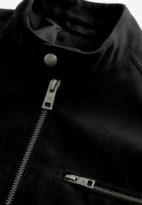 Schwarze Wildlederjacke mit hohem Kragen, ausgestattet mit einem Frontreißverschluss und einem seitlichen Reißverschlussfach, das silberne Beschläge und eine glatte Textur zeigt.