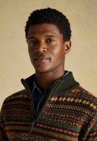 Zöld kötött pulóver magas gallérral, sárga, narancs és burgundi színű, sokszínű mintákkal. Cipzáras záródás és texturált anyag.