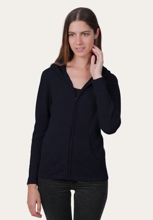 Sweat-shirt à glissière bleu marine en tissu doux, comprenant une fermeture éclair à l'avant, une capuche et des poches latérales. Porté avec un haut noir et un jean sombre.