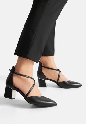 Černé kožené boty s nízkým podpatkem a šněrovacími pásky přes kotníky, nošené s černými zkrácenými kalhotami na bílém pozadí.