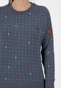 Niebieski sweter z dzianiny z białym wzorem kotwicy i kropek, różowym emblematu serca oraz brązową okrągłą łatą na rękawie.