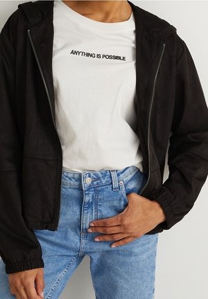 Person iført en hvid t-shirt med teksten "ALT ER MULIGT", sort jakke med lynlås og blå jeans med høj talje med en hånd hvilende på hoften.